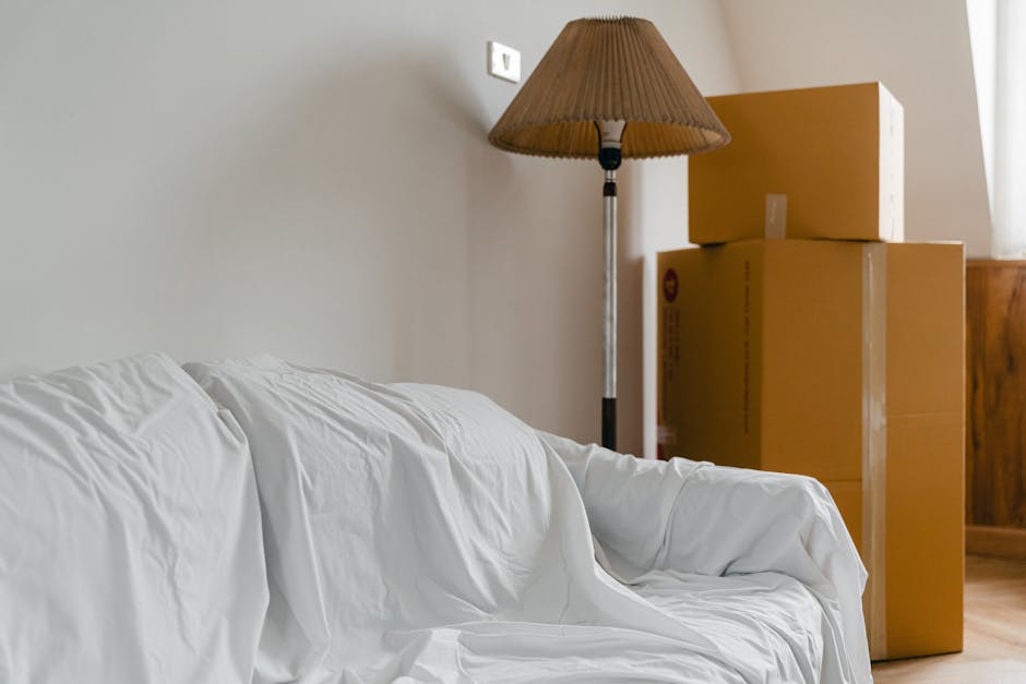 A room inside a residential property showing a professional moving process with furniture and moving boxes. In the foreground, several cardboard boxes of varying sizes are stacked on the polished wood