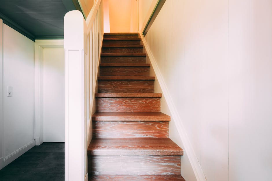 A top-down view of a narrow, spiral staircase with wooden handrails and black metal balustrades, descending into a dimly lit interior space. The staircase is carpeted with a runner, and the surroundin