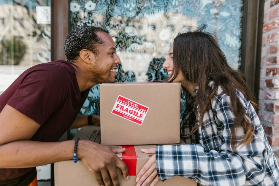 Two women, standing inside a room with a plain, textured grey wall, are surrounded by packed cardboard boxes of various sizes, some sealed with red tape. One woman is holding a medium-sized box with a