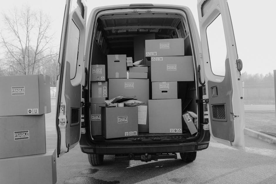 Photograph showing a professional home relocation process involving an interior room with a cardboard box and a stack of packed items on a wooden floor. The doorframe is visible leading to another par